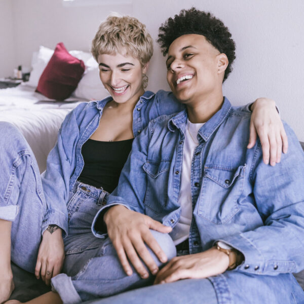 Two people sit up against a wall, touching each other and laughing, in a bedroom.